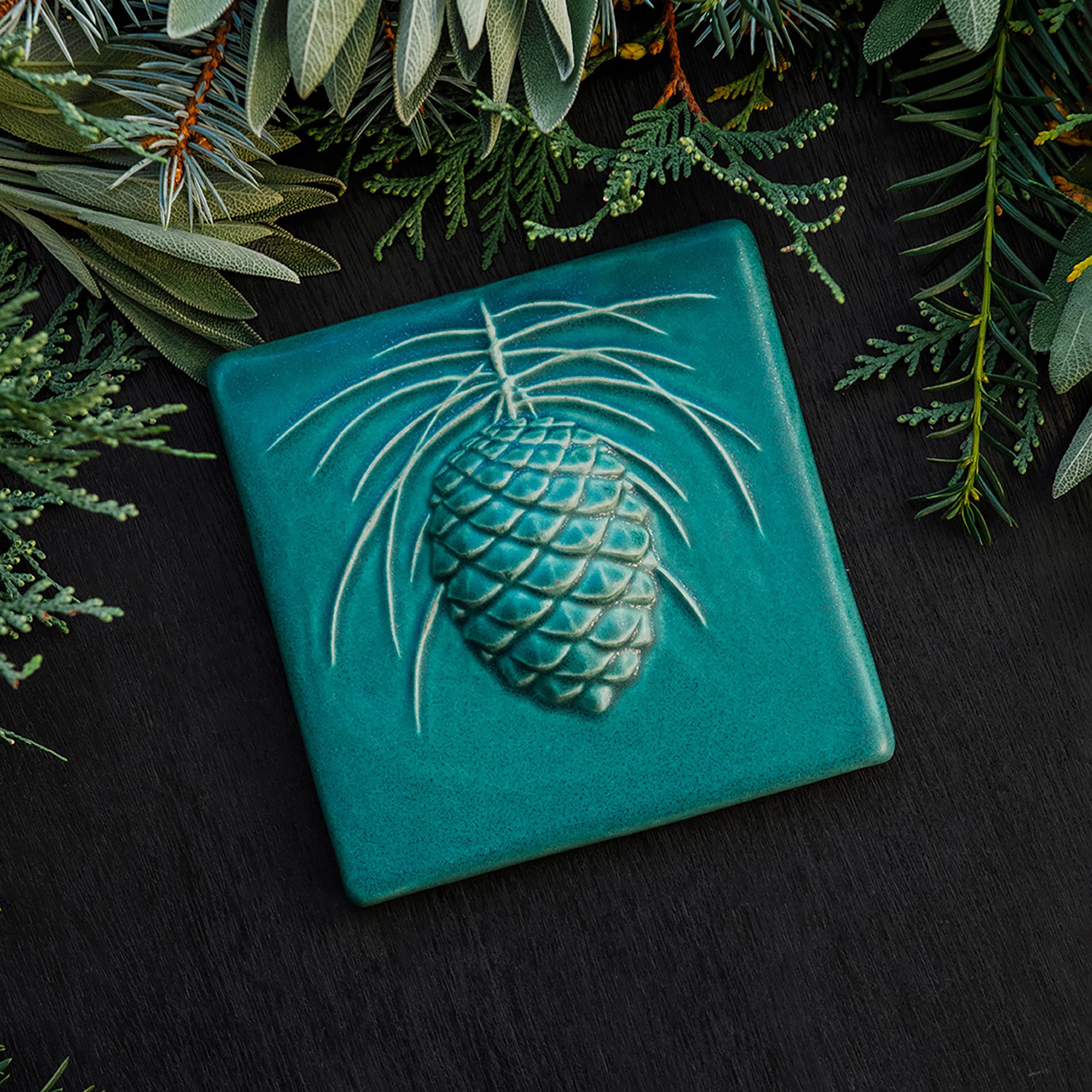 The 6x6 Pinecone tile features a large pinecone in the center of the tile with pine needles encircling it. This piece shown in Pewabic blue- a turquoise blue color.