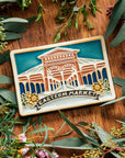 A hand-painted Eastern Market Postcard Tile rests on a wooden table with a warm finish surrounded by dainty wax flowers and seeded eucalyptus. 