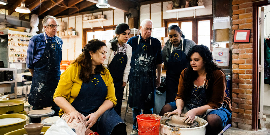 A Pewabic educator demonstrates a wheel throwing technique while five students stand around her to watch.