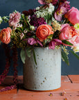 A Pewabic Crock featured in the icy-blue Frost glaze. Dark iron flecks are visible on the glossy surface of the vessel. The crock is set on terracotta roof tiles with a lime-wash backdrop and brilliant summer florals, including garden roses. 