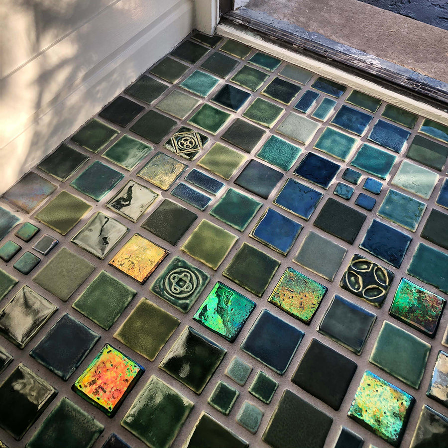 A blend of blue and green tiles in a range of textures and glazes on the floor at the  entryway of a home. The door is slightly ajar, welcoming you into the space. 