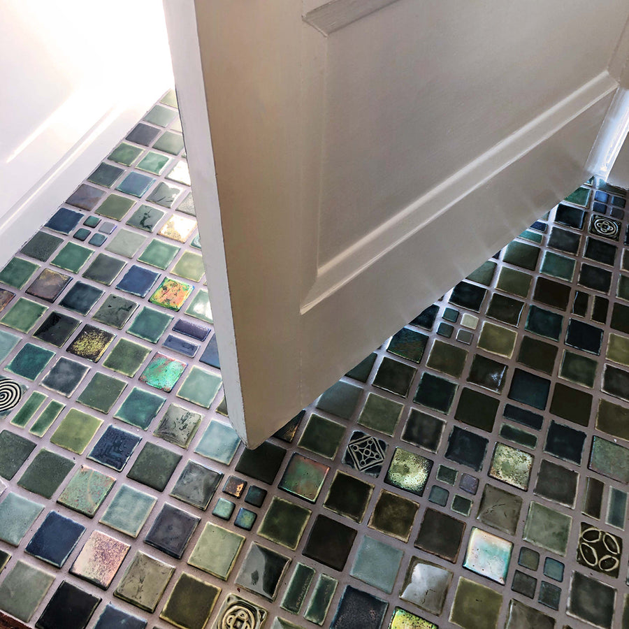 A blend of blue and green tiles in a range of textures and glazes on the floor at the  entryway of a home. The door is slightly ajar, welcoming you into the space. 