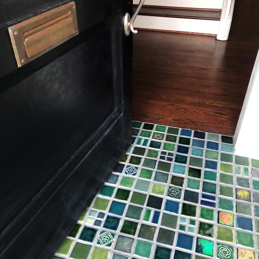 A blend of blue and green tiles in a range of textures and glazes on the floor at the  entryway of a home. The door is slightly ajar, welcoming you into the space. 