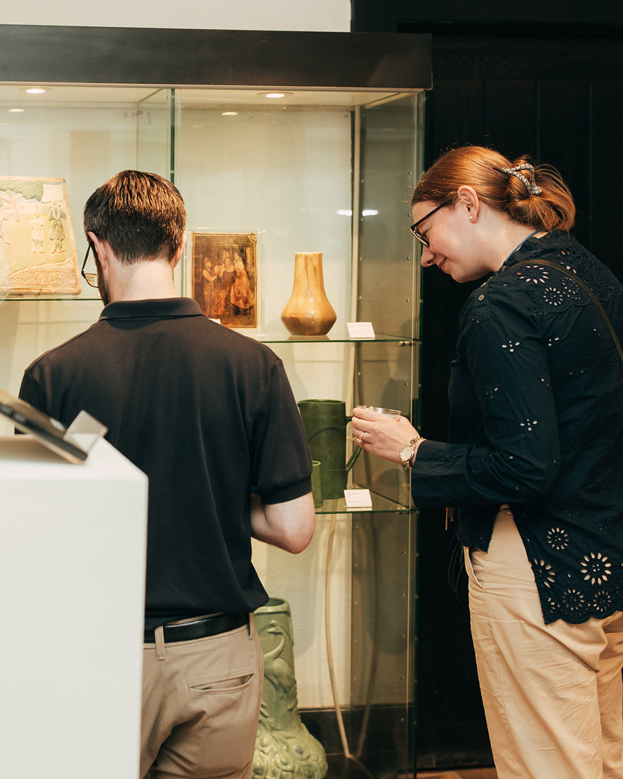 A man and a woman look stand in front of historic ceramic tiles and vessels behind glass in Pewabic's museum.