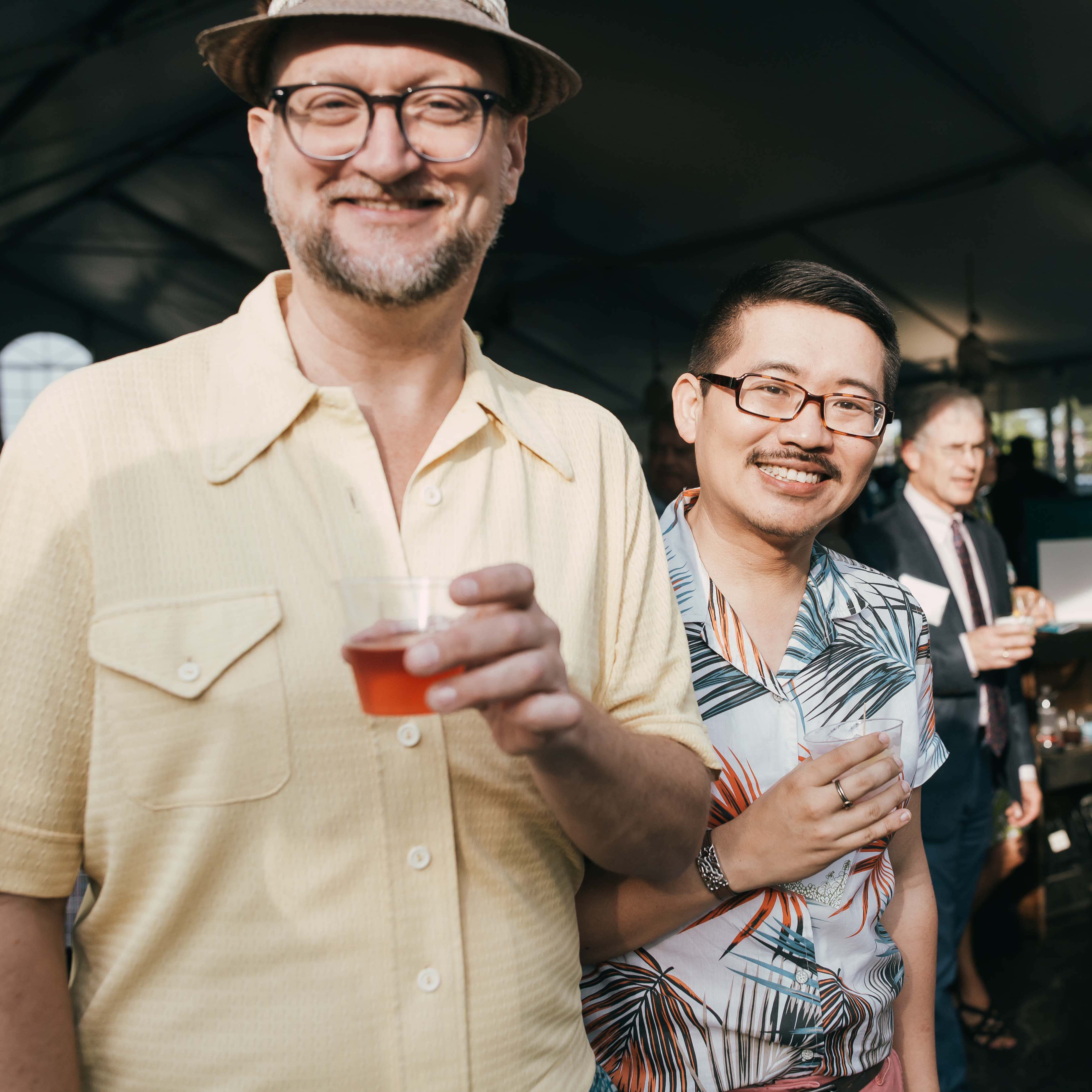 Two men smile with cocktails in hand during the Pewabic Preview Party Fundraiser. The man on the left has a butter yellow shirt and hat, while the man on the right sports a palm frond patterned button up shirt