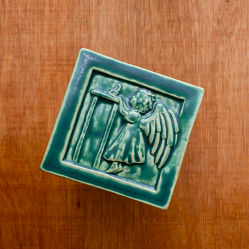 The Little Angel Girl Tile features a a toddler in a long nightgown with large feathered wings. She is on tip toes reaching for a lit candle on a high table. The candle is pulled towards her and the flame is wavering from the breeze created as she tries to blow out the flame. This tile features the matte blueish-green Pewabic Green glaze.