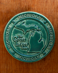 This round Michigan Trivet Tile features the outline of the state's two peninsulas including some of the larger northern islands of Michigan. To the left of the design reads "The Great Lakes State" and surrounding it, in a border, reads the word Michigan four times. 