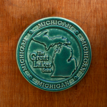 This round Michigan Trivet Tile features the outline of the state's two peninsulas including some of the larger northern islands of Michigan. To the left of the design reads "The Great Lakes State" and surrounding it, in a border, reads the word Michigan four times. 
