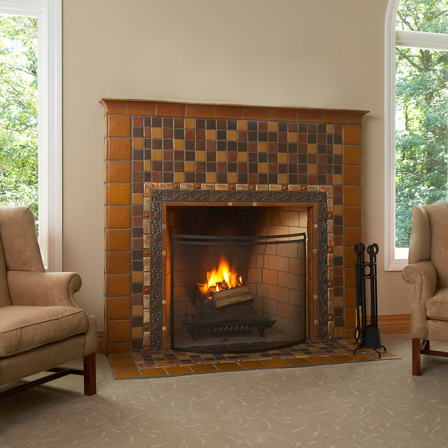 Lit fireplace in the center of two beige couches. The tile is rustic and comprised of a blend of brown and earth-tone tiles. A tasteful Iridescent custom-cut trim lines the border alongside brown tiles in an acorn motif.