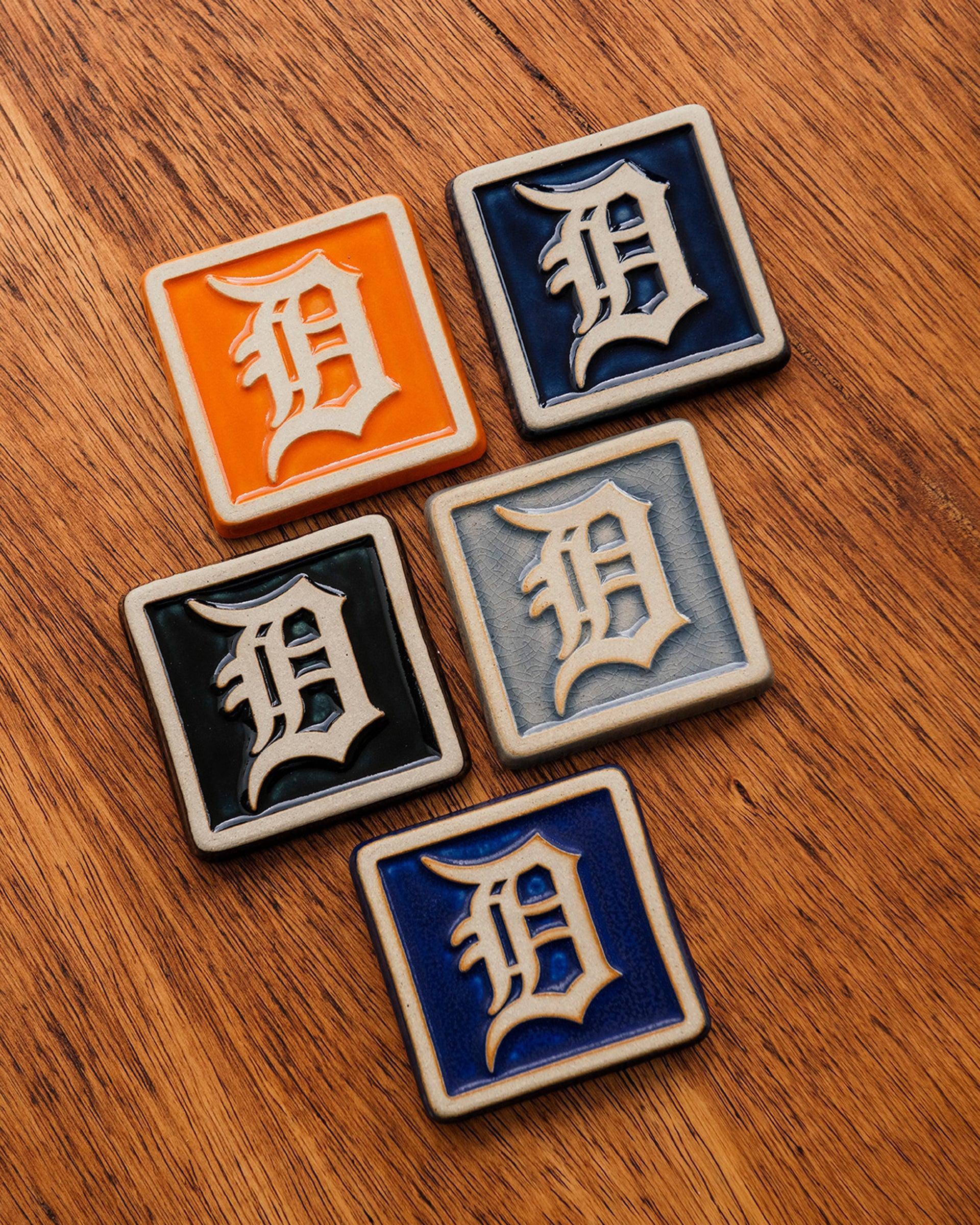 Four Old English 'D' logo tiles in a range of colorful glaze options on a wooden background.