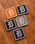 Four Old English 'D' logo tiles in a range of colorful glaze options on a wooden background.