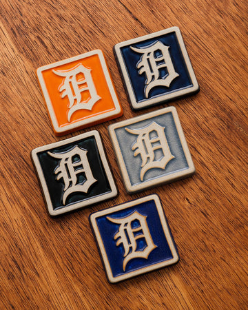 Four Old English 'D' logo tiles in a range of colorful glaze options on a wooden background.