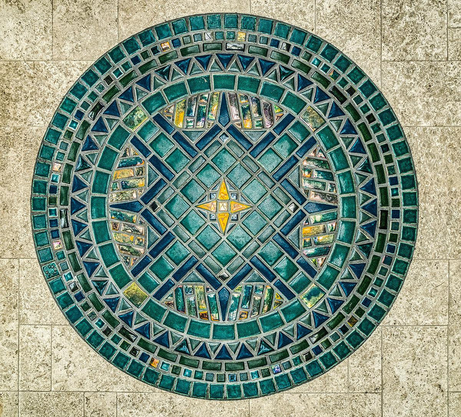 An aerial view of a custom, circular, tile medallion with elements of a classic compass. The design incorporates glossy, matte, and iridescent glazes in an intricate geometric pattern.  