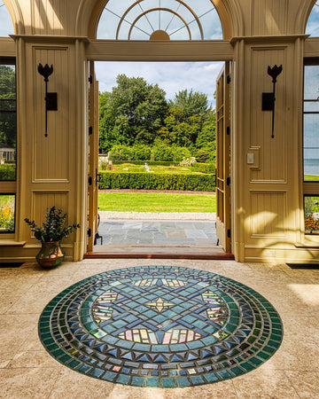 A custom, circular, tile medallion is the focal point of this photo which includes large doors opening to an impressive garden view. The tile design has elements of a compass and is rich in glossy blue and green glazes paired with a stark Aurora Iridescent glaze.