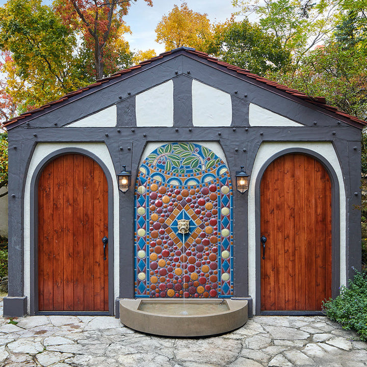A tile mural full of leaves, berries, circles, and other shapes is installed on the side of a out building with two large wooden doors flanking the design. A fountain of water comes out of the tile installation and into a cement pool at its base.