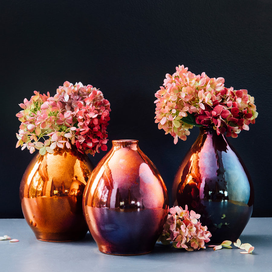 Three Copper glazed Teardrop vases stand filled with bushy hydrangea stems. The glaze on these vases have a rosy, purple and blue variation.