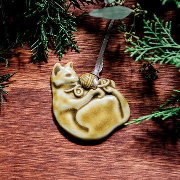 The ceramic Cat with Yarn Ornament features a cat on its back with its paws grasping a ball of yarn. Its face is happy and playful. Both the cat and the yarn are featured in our matte yellow-cream Wheat glaze.