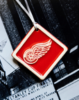 The Red Wings Ornament is a bright red diamond shaped tile. The Red Wings logo is raised in its center and has been scraped of glaze giving it a white appearance. There is a simple line border around the tile that is also scraped white.
