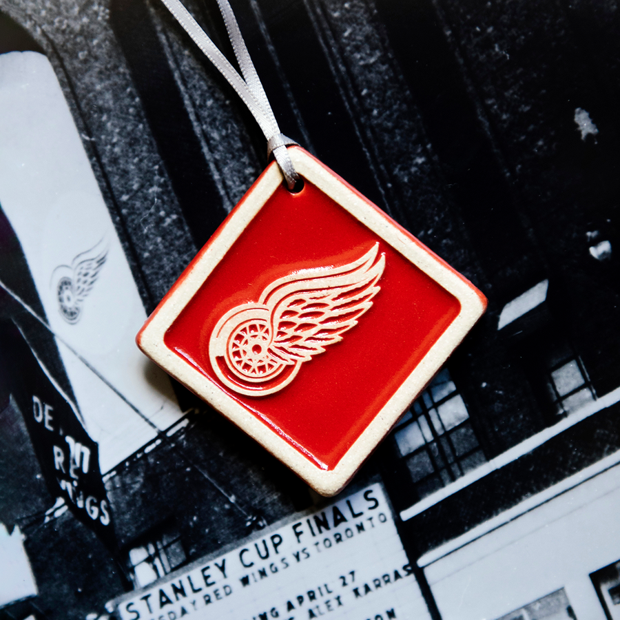 The Red Wings Ornament is a bright red diamond shaped tile. The Red Wings logo is raised in its center and has been scraped of glaze giving it a white appearance. There is a simple line border around the tile that is also scraped white.