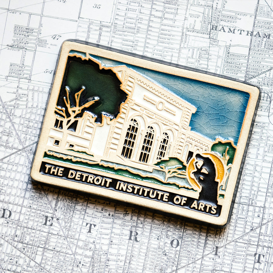 The bright white building of the Detroit Institute of Arts is featured in this hand painted tile. Large bushy trees frame the facade with the dark outline of the Thinker statue in front of it. Under the building design are the words "The Detroit Institute of Arts".