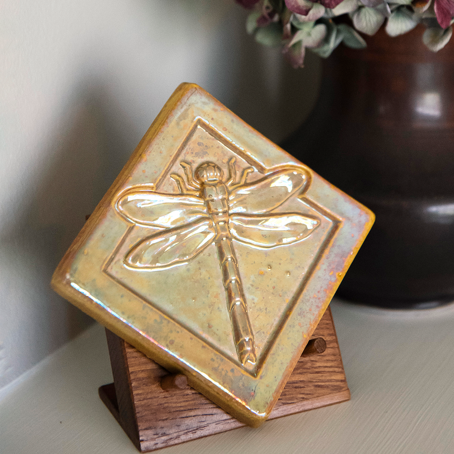 The Dragonfly Tile features a detailed dragonfly with wings outstretched. It is sitting horizontally on the square tile with a thick border around the edge. The pinkish gold metallic Blush Iridescent glaze features lots many variations. Depending on the lighting in the room, these pieces will pick up different hues.