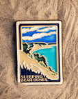 The Sleeping Bear Dunes Tile features the serene Lake Michigan and the sandy beaches and dunes covered on top by bushes and greenery. The words "Sleeping Bear Dunes" line the bottom of the design. The hand painted tile boasts blues and greens and pale yellow sand.