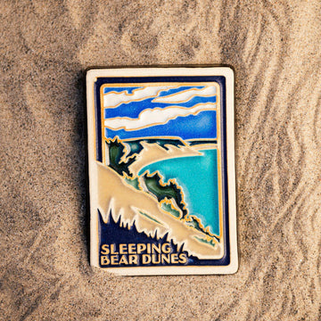 The Sleeping Bear Dunes Tile features the serene Lake Michigan and the sandy beaches and dunes covered on top by bushes and greenery. The words "Sleeping Bear Dunes" line the bottom of the design. The hand painted tile boasts blues and greens and pale yellow sand.