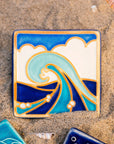 The Wave Tile features a line drawing of a large, curled wave. There are bubbles scattered around its base with a huge puffy cloud behind it that almost covers the bright blue sky. The hand painted tile has multiple blue hues with bright white accents.