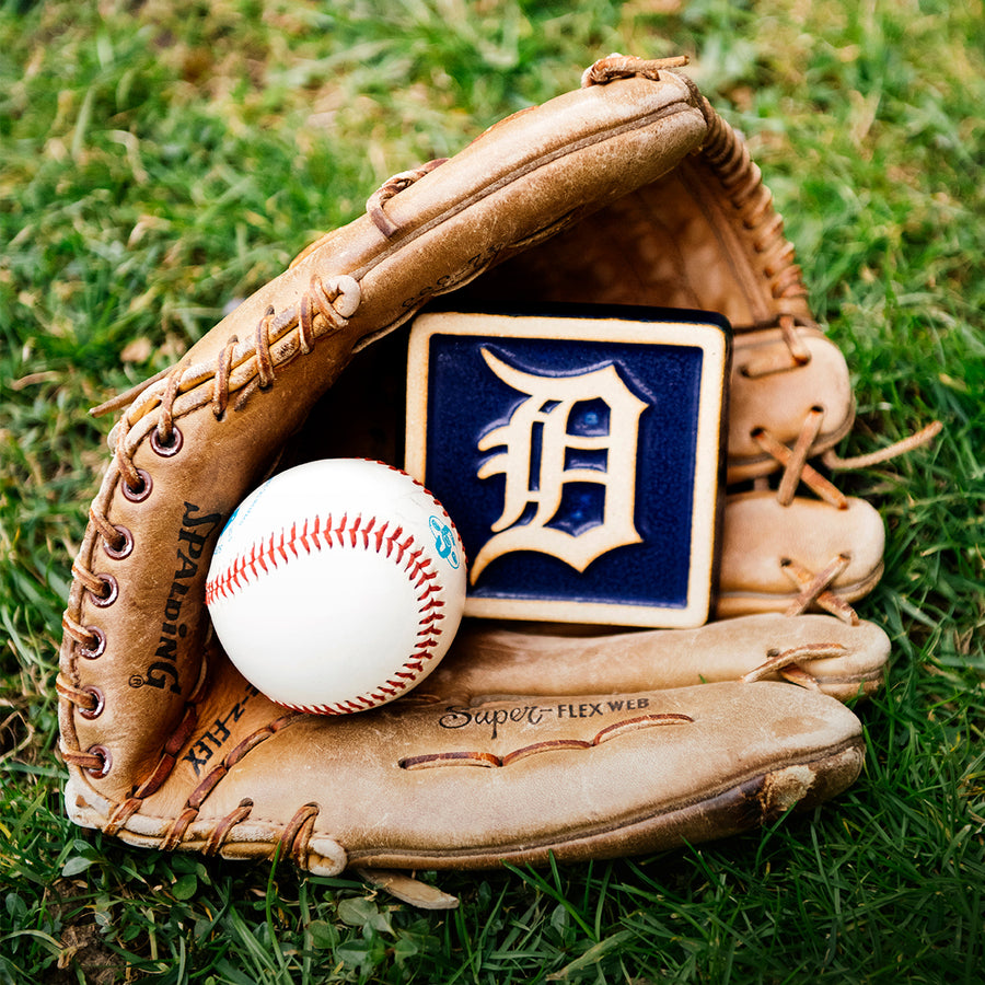 A leather baseball mitt is nestled in bright, green grass. Inside the mitt lays a baseball and our 4x4" Tigers Old English "D" Tile in a Navy Blue and light   "Natural" clay border. The Old English "D" logo is also outfitted in the light clay body, allowing it to stand out against its bright backdrop.
