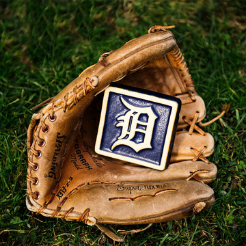 A leather baseball mitt is nestled in bright, green grass. Inside the mitt lays a our 4x4" Tigers Old English "D" Tile in a Navy Blue and light "Natural" clay border. The Old English "D" logo is also outfitted in the light clay body, allowing it to stand out against its bright backdrop.
