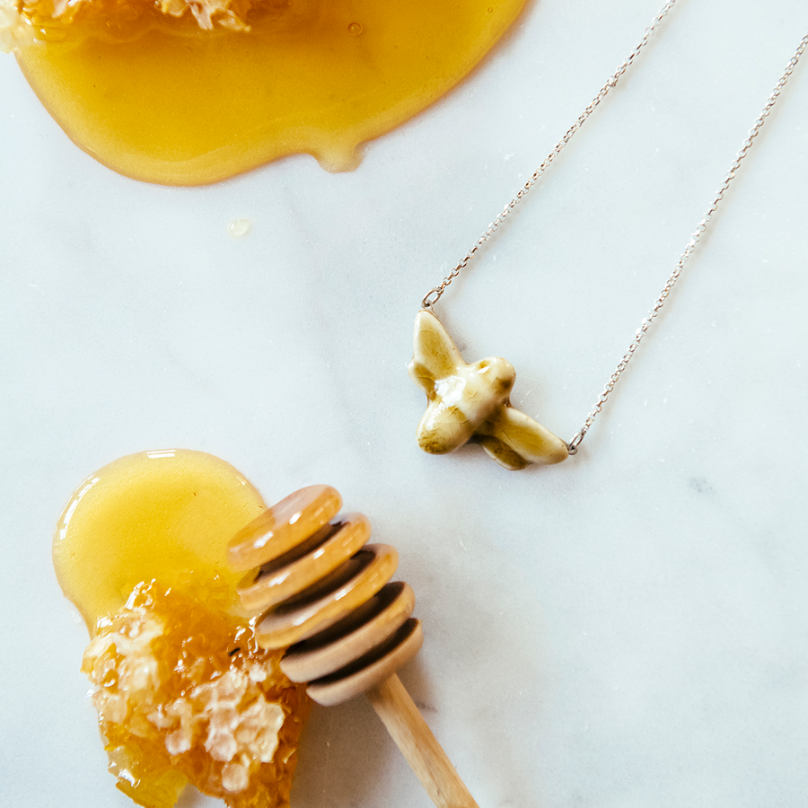 The Honey Gloss bee pendant is attached to a silver chain and sits next to a honey dipper.