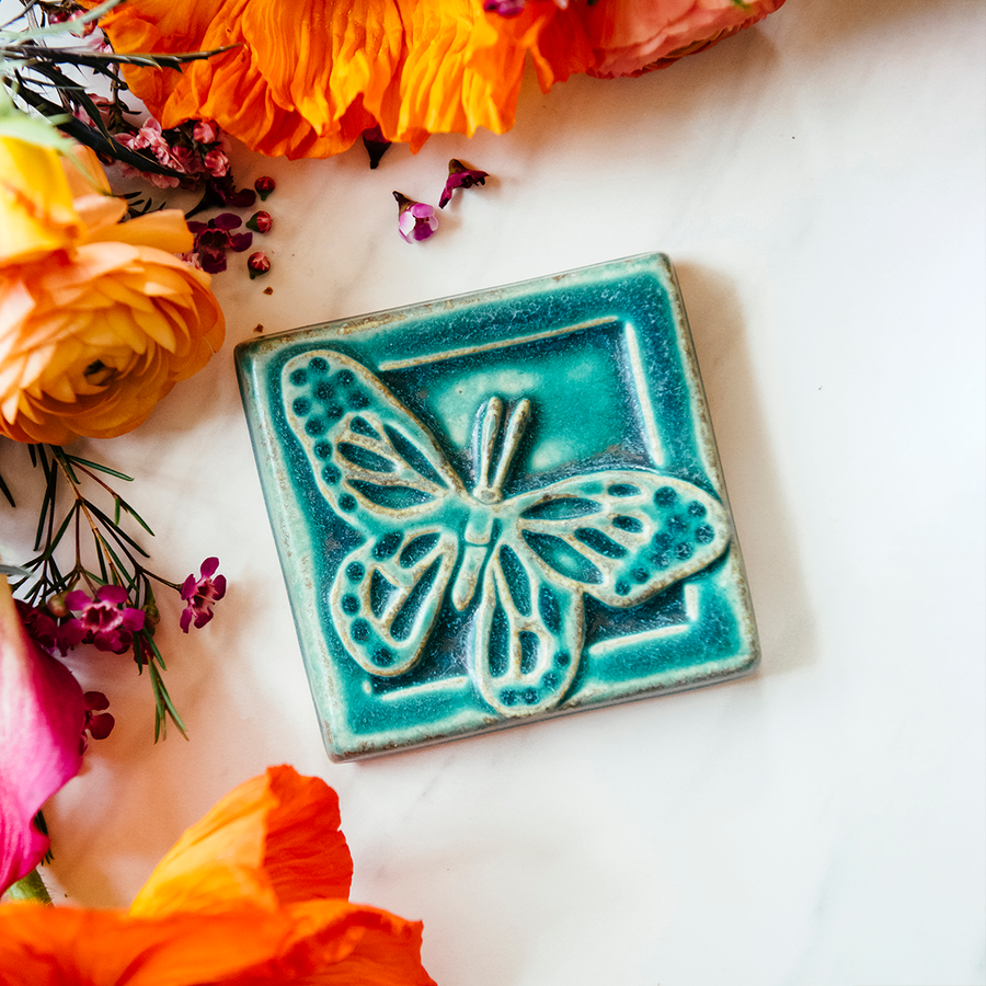 The ceramic Butterfly tile's design sits diagonally on the 4x4 square tile. Its wings are outstretched and it is floating above a depressed border that surrounds the tile. This gives the illusion that the butterfly is flying out toward you. This tile features the matte turquoise Pewabic Blue glaze.