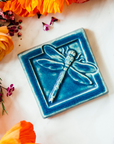 The Dragonfly Tile features a detailed dragonfly with wings outstretched. It is sitting horizontally on the square tile with a thick border around the edge. This Dragonfly Tile features the matte french blue Peacock glaze.