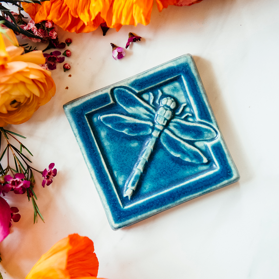 The Dragonfly Tile features a detailed dragonfly with wings outstretched. It is sitting horizontally on the square tile with a thick border around the edge. This Dragonfly Tile features the matte french blue Peacock glaze.
