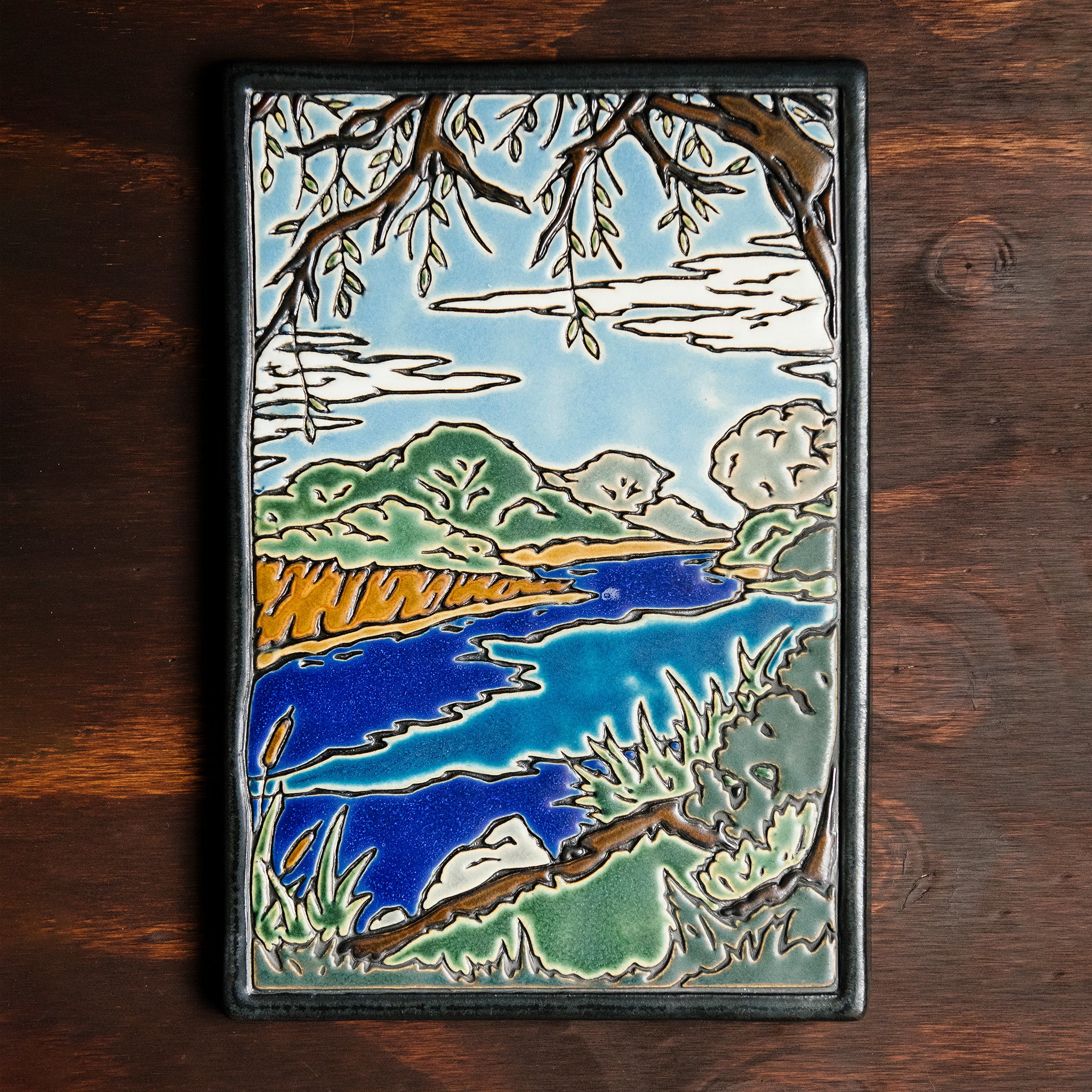 The Lake Okonoka Tile features a snapshot of the lake with bushes lining its banks and a large willow tree in the foreground that frames it. This gives the impression that you are seated on the grass under the tree next to the serene water. This tile is hand painted in blues and greens.