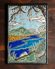 The Lake Okonoka Tile features a snapshot of the lake with bushes lining its banks and a large willow tree in the foreground that frames it. This gives the impression that you are seated on the grass under the tree next to the serene water. This tile is hand painted in blues and greens.