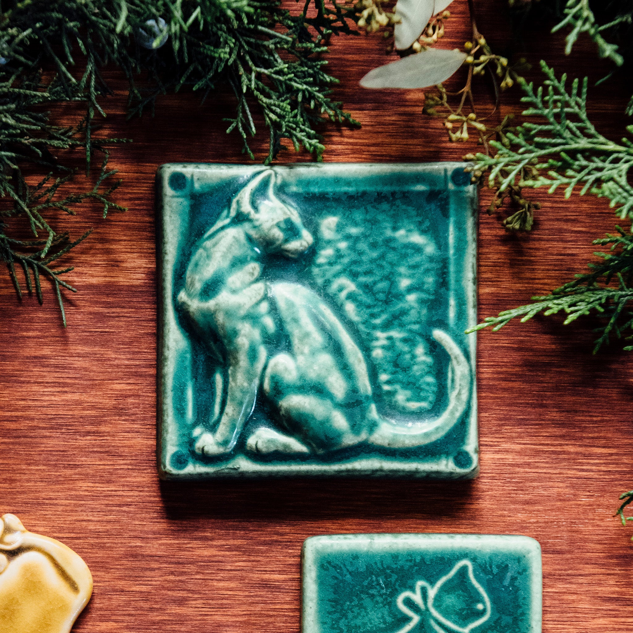 A tile sits on a wooden table surrounded by greenery. The matte green, Pewabic Green embossed tile shows a sleek cat sitting up straight with its head turned backward towards its flicking tail.