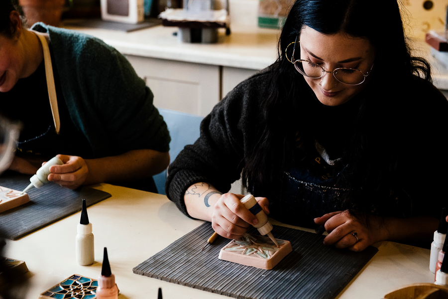 A young woman sits at a table. She uses a small bottle with a pointed tip to squeeze liquid glaze into the crevices of a patterned tile.