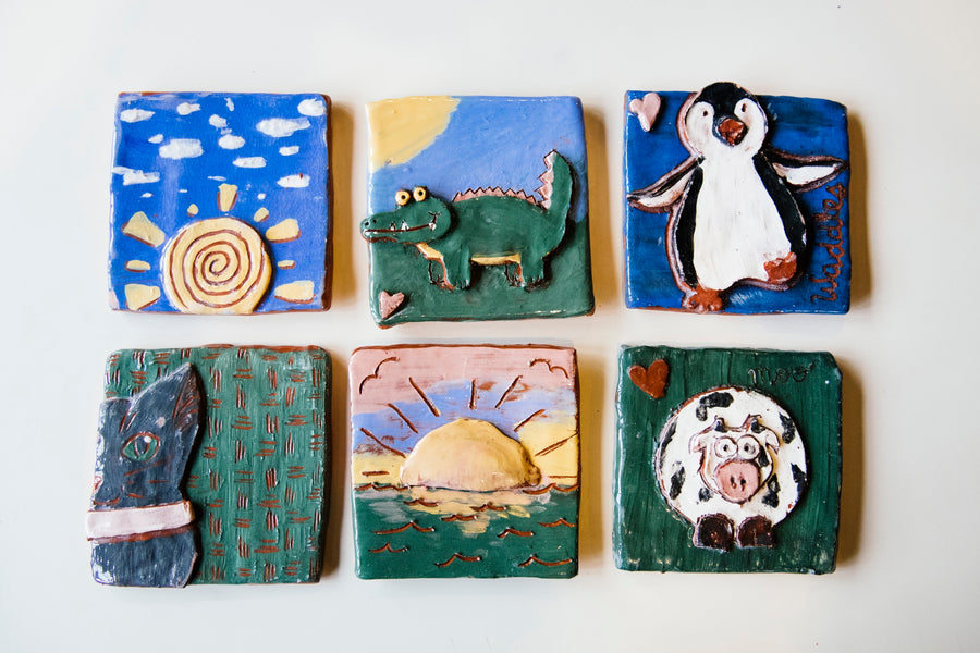 Six tiles are layed out on a white table. The tiles have been carved to have designs featuring animals and landscapes. They have been painted with colored slip.