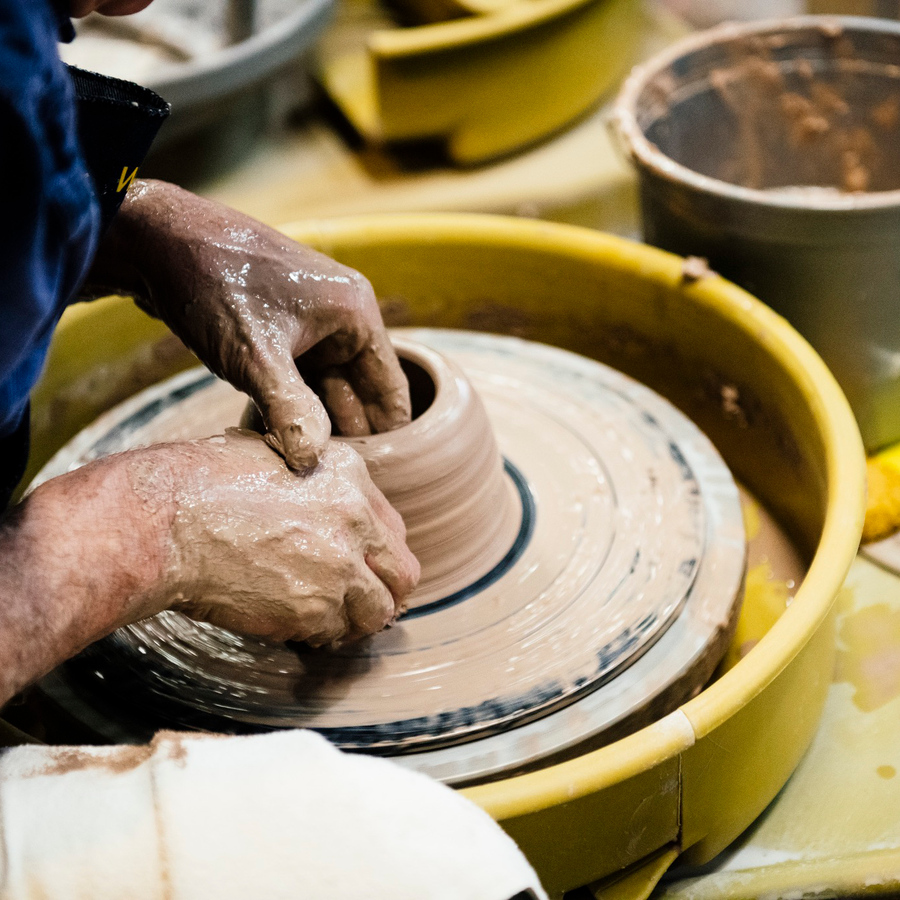 Two clay-covered hands are molding a cup out of wet clay on a throwing wheel.