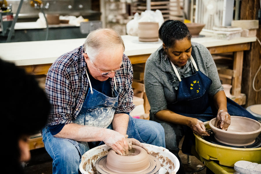 Two students sit side by side, each molding clay on their own pottery wheel in our education studio.