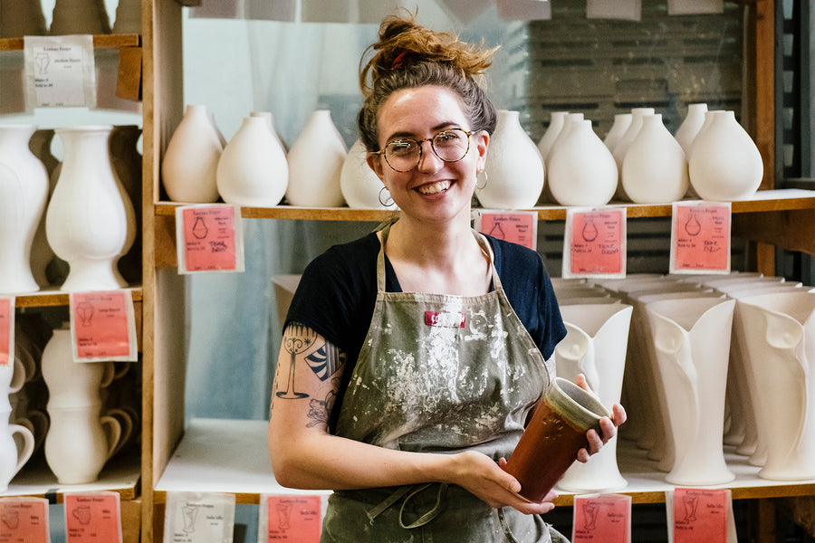 A Pewabic vessel maker sits in front of shelves filled with the wheel thrown vases she has created. Her be-speckled face smiles at the camera as she holds a cinnamon colored Pewabic pint. 