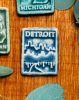 The Detroit Magnet is a rectangular tile featuring a tree lined road leading towards the Detroit skyline. The word Detroit is in bold letters across the top of the tile.