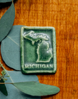 The Michigan Magnet features the state's two peninsulas with the word "Michigan" written below them. This piece has a simple border around the entire design.