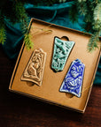 Three ceramic ornaments are sitting in a gift box and placed under a Christmas tree.