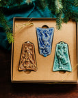 Three ceramic ornaments are sitting in a gift box and placed under a Christmas tree.