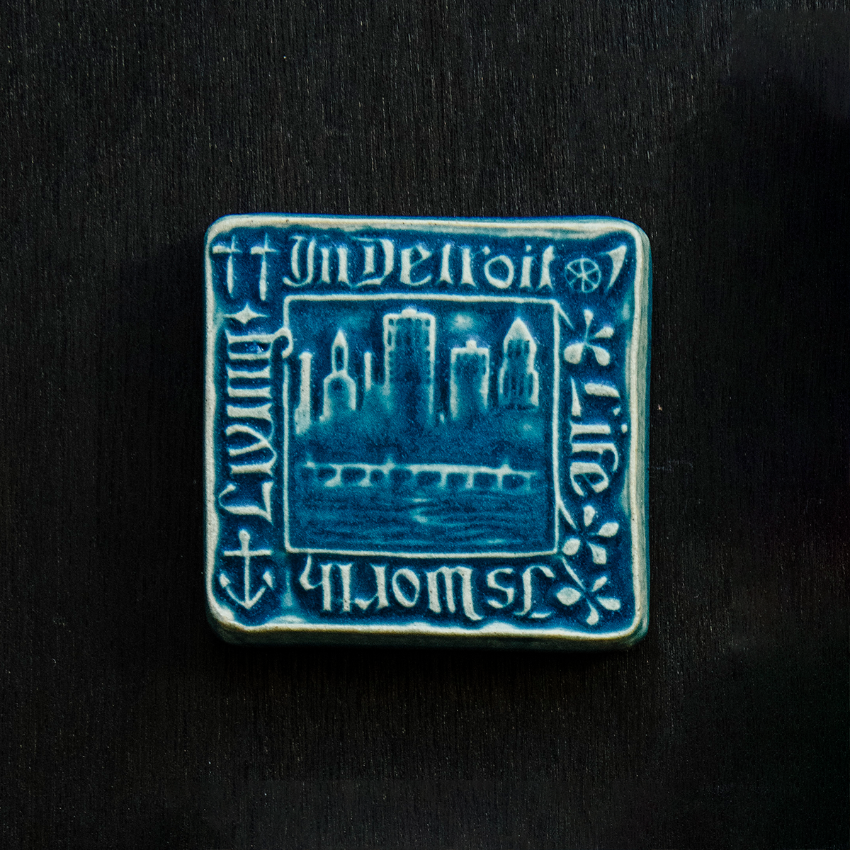 The Old Detroit Tile features an image of the Detroit skyline circa 1906 with the Belle Isle Bridge and the waters of the Detroit river in the foreground. A thick border around the design says "In Detroit Life is Worth Living" with a small symbol of four Detroit industries in each corner.