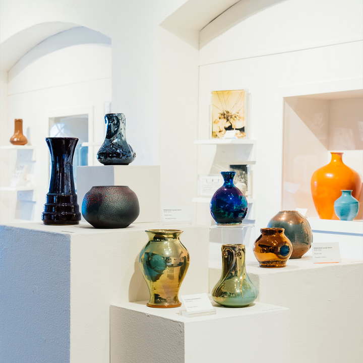 A display of test iridescent glazes sits in our museum space. The shiny metallic colors ranges from gold to blue to copper to orange. 