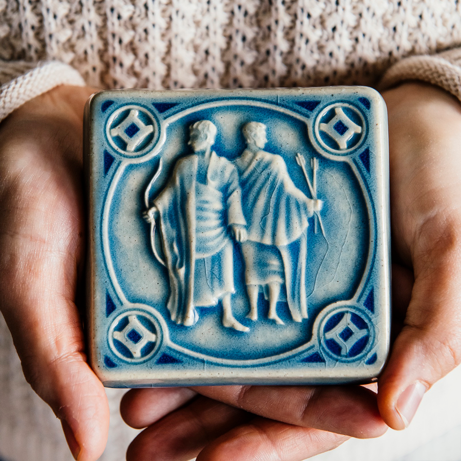 The glossy pale blue Celestite glaze pools across the low-relief sculpted twin forms of this Gemini tile. Two warm hands hold the tile in natural light to show off the glossy surface.