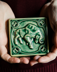 The glossy green Emerald glaze pools across the low-relief sculpted bull form of this Taurus tile. Two graceful hands hold the tile in natural light to show off the glossy surface.